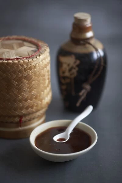 Bowl containing plum sauce and spoon, rice basket and bottle in background