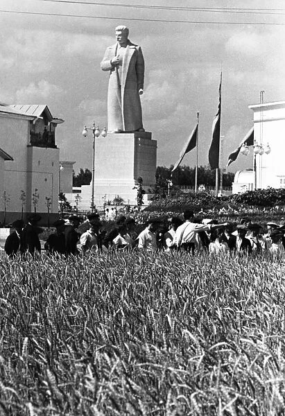 The all-union agricultural exhibition in moscow, august 1939, visitors to the exhibition examining specimens of a couch-grass  /  wheat hybrid (developed by member of the academy of sciences, tsitsin) at the demonstration field of the grain pavillion, in the background is a newly erected statue of stalin