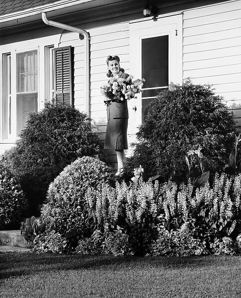 Woman standing on front porch of house holding bunch of flowers, (B&W)