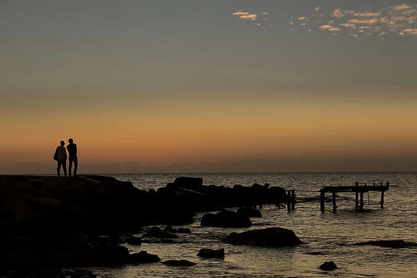 Silhouette Of Two People Standing At The Waters Edge Watching The Sunset Over The Water; Paphos, Cyprus