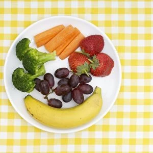 Banana, broccoli, carrot sticks, strawberries and grapes on plate, on yellow checked tablecloth