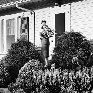 Woman standing on front porch of house holding bunch of flowers, (B&W)